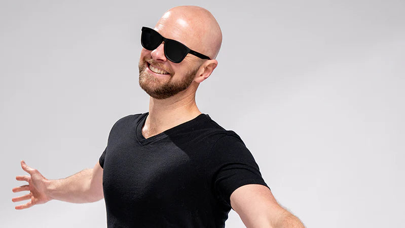 Smiling person wearing black sunglasses and a black shirt, posing with arms outstretched against a light background.