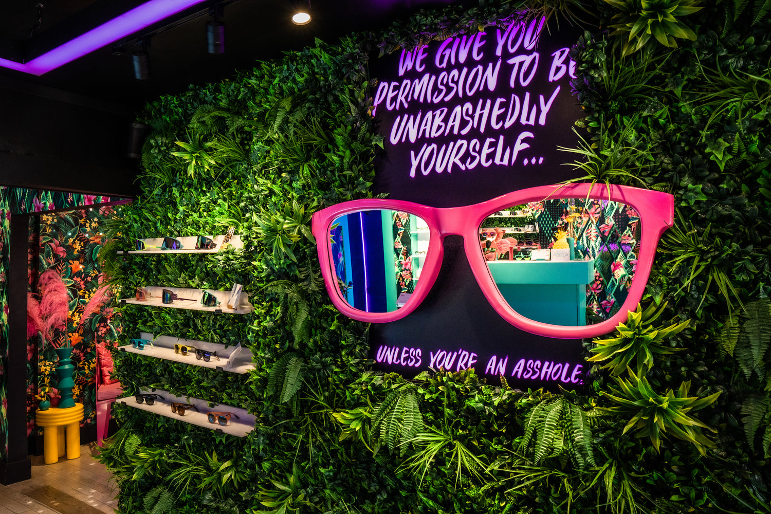 A vibrant store display with neon lights, oversized pink glasses, and a cheeky sign amidst lush greenery.