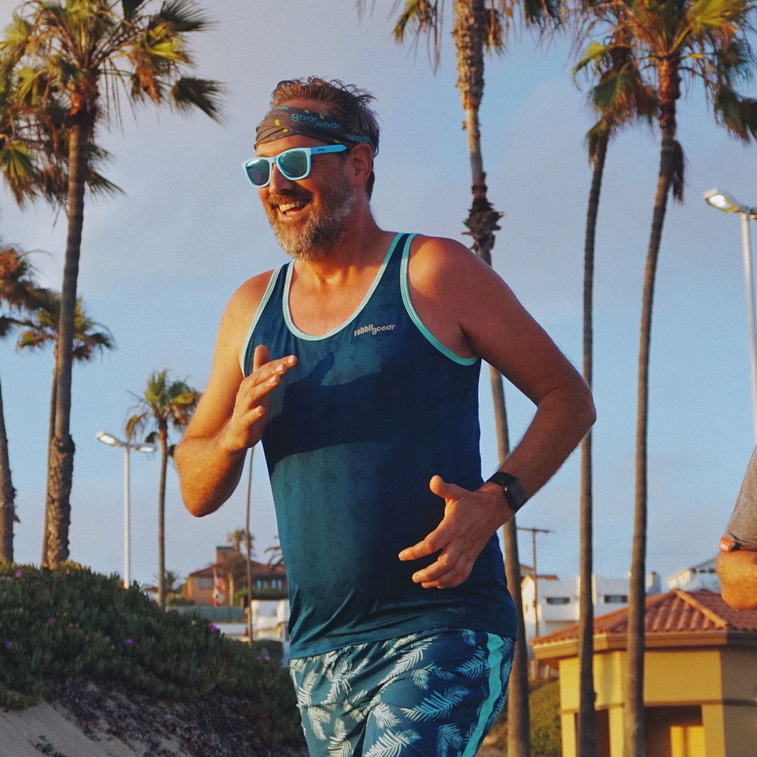 A man in a blue tank top, shorts, and bandana running, wearing blue square shaped sunglasses
