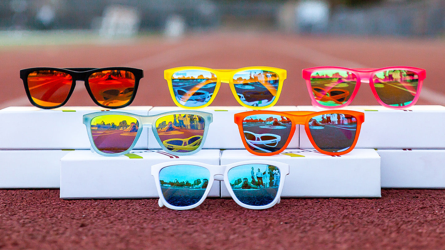 A colorful display of six pairs of sunglasses, each with reflective lenses, arranged in three rows on white boxes. The sunglasses frames come in various bright colors