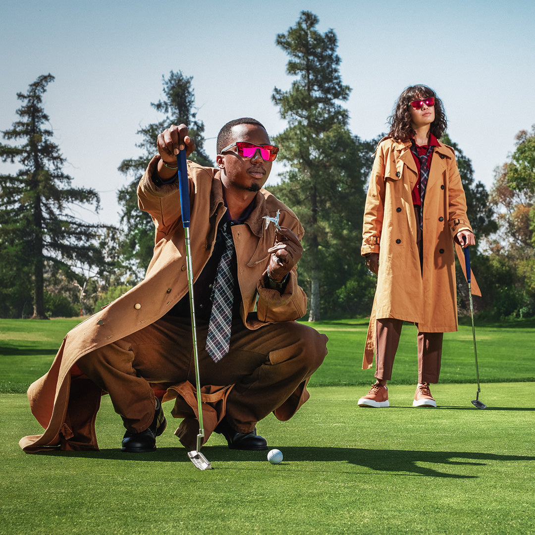 Two people wearing long tan trench coats and red sunglasses are on a golf course. One person is squatting, aiming a putt while holding a trophy, while the other stands in the background with a golf club.