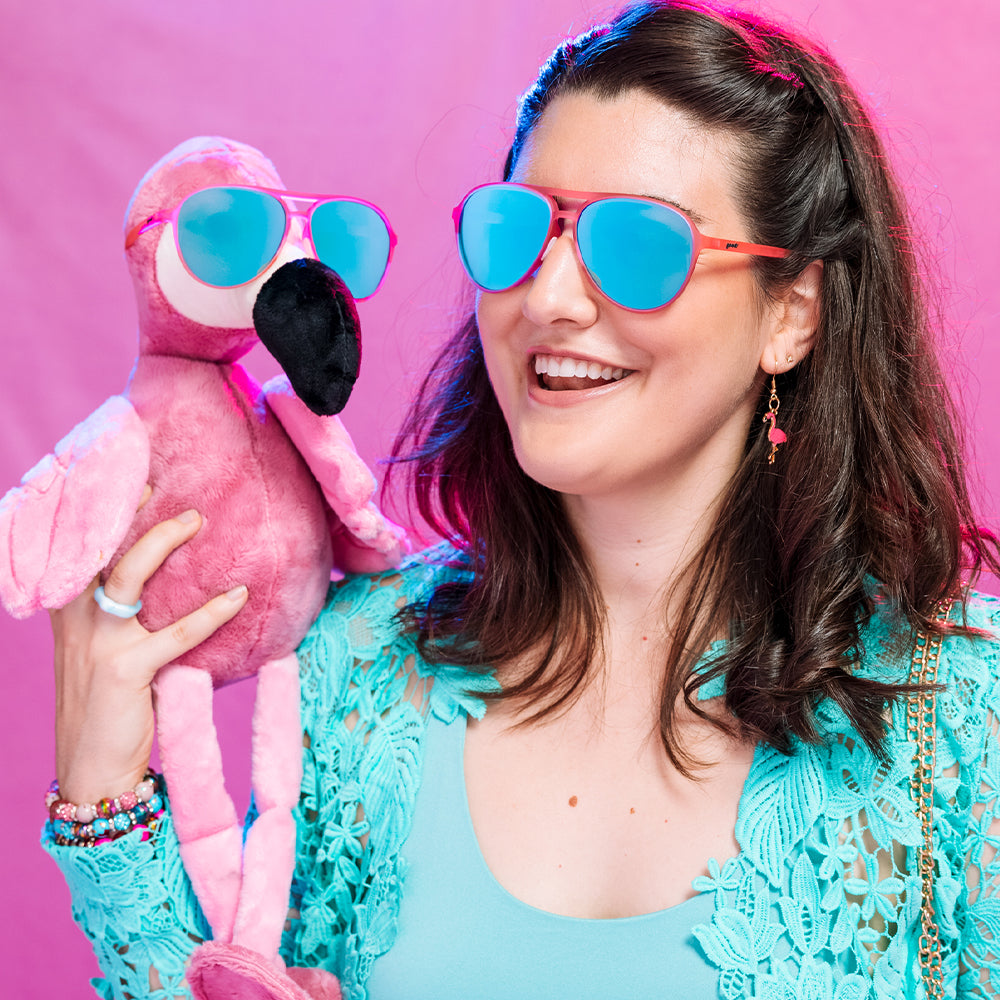 Person in a turquoise top holding a plush flamingo with sunglasses against a pink background.